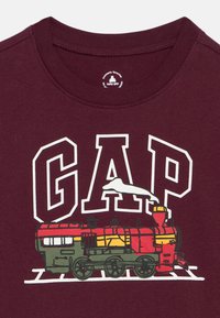 Παιδικό πουκάμισο μπορντό με μεγάλα λευκά γράμματα GAP και πολύχρωμο γραφικό παλαιού ατμοκίνητου τρένου που διασχίζει σιδηροδρομικές γραμμές.