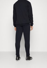 Person som bär svarta träningsbyxor, svart sweatshirt och svarta Adidas Predator fotbollsskor står mot en enkel vit bakgrund.