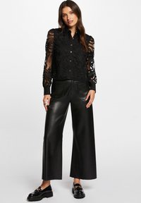 Blusa nera in pizzo con motivi floreali e chiusura a bottoni, abbinata a pantaloni a gamba larga in pelle nera e mocassini neri lucidi.
