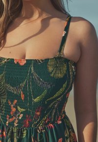 Rochie florală cu un decolteu ajustat, modelat, în verde, cu modele florale colorate și bretele subțiri, ajustabile. Țesătură moale, texturată.