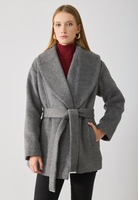 Manteau en laine gris avec un col oversize, ceinture nouée à la taille, poches latérales et texture douce. Porté par-dessus un col roulé bordeaux.
