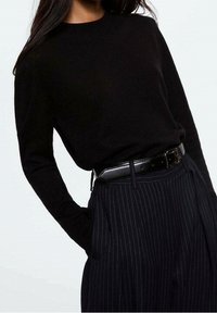 Pull en maille noire avec col rond, associé à un pantalon taille haute à rayures fines foncées et une ceinture noire brillante. Tissu texturé et coupe ajustée.