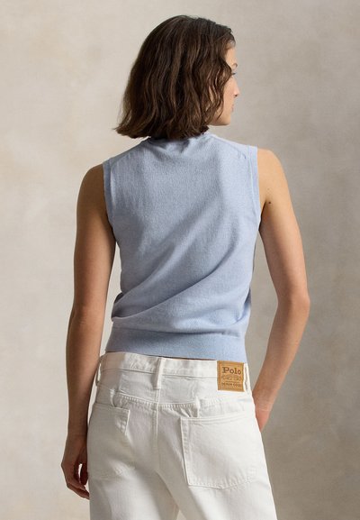 Γυναίκα με καστανά μαλλιά μέχρι τους ώμους που φοράει αμάνικο ανοιχτό μπλε τοπ και άσπρο τζιν, γυρισμένη από πίσω σε μπεζ φόντο.