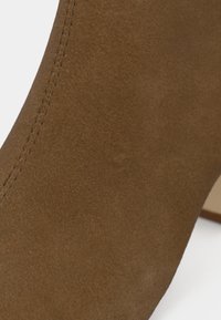 Close-up de bota de tornozelo em camurça castanha com costuras visíveis e salto bloco contra um fundo branco.
