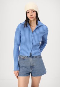 Cardigan en maille bleu clair à côtes, à manches longues et col, comportant cinq boutons. Assorti à un short en denim effiloché. Bonnet en tricot beige.