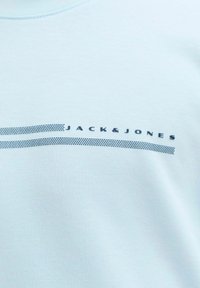 Ανοιχτό μπλε ύφασμα με μικρό σκούρο μπλε κείμενο "JACK & JONES" πάνω από δύο παράλληλες τεθλασμένες γραμμές στο κέντρο.