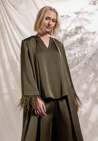 Tenue vert olive comprenant un haut ample à encolure en V avec des accents de plumes sur les manches, associé à un pantalon large assorti. Texture lisse.