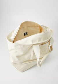 Lys beige tote bag med glidelås, frontlomme og logomerke inni. Myk tekstur og slitesterkt stoff. Strukturert form med korte håndtak.