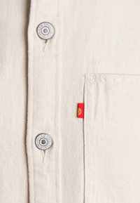 Camicia color crema con bottoni a pressione, dotata di bottoni in metallo e un'etichetta logata rossa. Tessuto testurizzato e dettaglio della tasca sul petto.