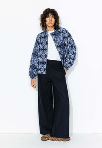 Veste bomber à motifs bleus avec un design en diamants, t-shirt blanc, pantalon noir ample et chaussures à enfiler à imprimé animal.