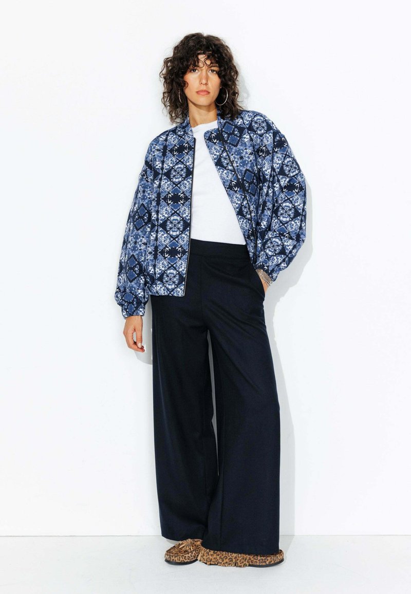 Veste bomber à motifs bleus avec un design en diamants, t-shirt blanc, pantalon noir ample et chaussures à enfiler à imprimé animal.