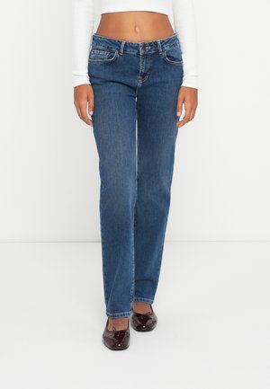 Woman wearing mid-rise blue straight-leg jeans, white long-sleeve cropped top, and dark red ballet flats standing against a white background.