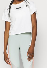 Camiseta blanca corta con logo negro "PUMA", acompañada de leggings verde claro con franjas laterales naranjas y cintura con cordón.