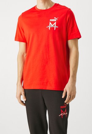 Homme portant un t-shirt rouge avec le logo Puma AC Milan et un pantalon noir avec le logo Puma AC Milan assorti sur la cuisse, debout devant un fond uni.