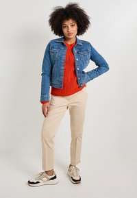 Chaqueta de mezclilla con dos bolsillos en el pecho, color azul, combinada con un suéter de punto naranja y pantalones ajustados beige. Zapatillas beige y negras.