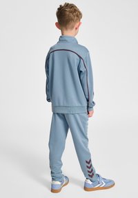 Niebieski dres sportowy z zapinaną na zamek kurtką i dopasowanymi spodniami. Czerwone akcenty i wzory w zygzak na spodniach. Miękki, lekki materiał.