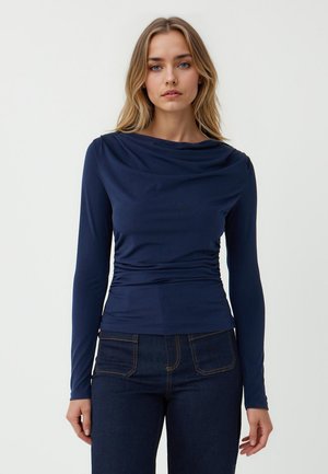 Top navy a maniche lunghe con collo a drappeggio, silhouette aderente e tessuto texture. Abbinato a jeans scuri a vita alta.