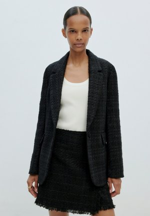 Zwarte gestructureerde blazer met een enkele knoop, gecombineerd met een bijpassende rok met franjes. Het binnenste topje is een aansluitende witte tanktop.
