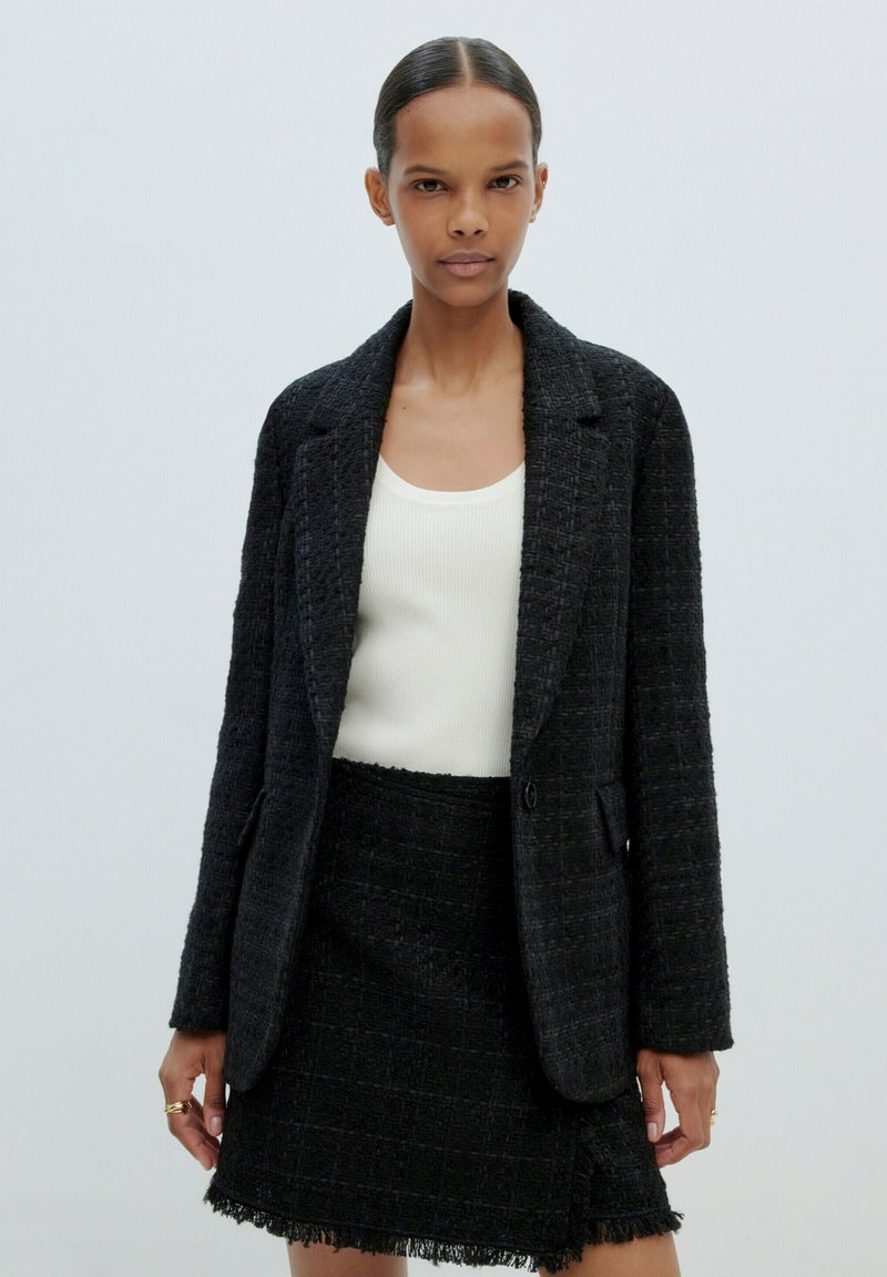 Schwarzer strukturierten Blazer mit einem einzelnen Knopf, kombiniert mit einem passenden Fransenrock. Das Innentop ist ein eng anliegendes weißes Tanktop.