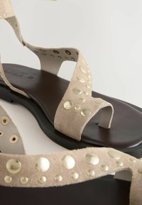 Beige suède sandalen met brede banden versierd met grote gouden studs, rustend op een donkerbruine rubberen zool.