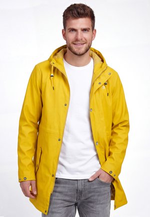 Man die glimlacht en een gele regenjas met capuchon, wit shirt en grijze spijkerbroek draagt, staand met één hand in zijn zak tegen een witte achtergrond.