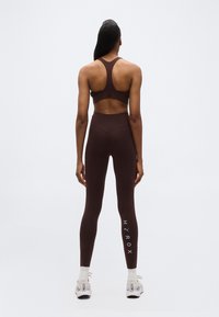 Γυναίκα π ου φοράει αθλητικό σουτιέν σκούρου καφέ χρώματος και κολάν με το λογότυπο "HYROX" στο αριστερό πόδι, στέκεται με την πλάτη προς τα εμπρός σε απλό φωτεινό φόντο.
