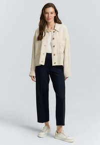 Beige cropped jakke i åndbart stof med to forlommer og knaplukning. Kombineret med marineblå vidde bukser og hvide sneakers.