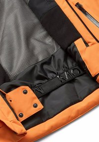 Binnenkant van een oranje jas met zwarte stof, een gestructureerde grijze voering, ritsen en een zwarte naamtagband. Inclusief drukknoop- en knoopdetails.