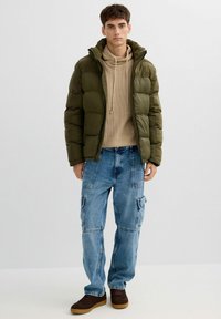 Chaqueta acolchada verde con capucha, suéter de punto color beige y vaqueros cargo azules. Zapatillas marrones. Materiales texturizados y ajuste relajado.