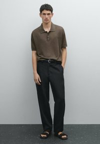 Hnědá polo košile s límečkem, doplněná černými pruhovanými kalhotami a černými sandály. Tkanina vypadá hladce a má uvolněný střih.