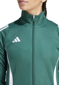 Giacca sportiva verde realizzata in tessuto liscio, presenta un colletto alto, chiusura con zip e tre strisce bianche sulle spalle, marchiata con un logo bianco.