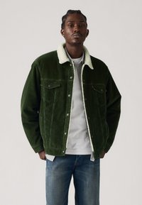 Green corduroy jacket with a white fur collar, featuring snap buttons and two front pockets, worn over a gray shirt and blue jeans.
