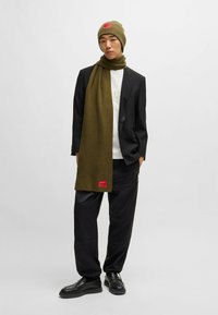 Beanie in maglia verde oliva e sciarpa coordinata con una finitura testurizzata. La sciarpa presenta un patch brandizzato rosso; indossato con un blazer e pantaloni neri.