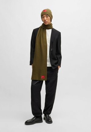 Gorro de punto en verde oliva y bufanda a juego con un acabado texturizado. La bufanda presenta un parche de marca en rojo; combinado con un blazer y pantalones negros.