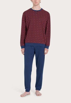 Uomo che indossa una maglietta bordeaux a maniche lunghe con piccoli motivi blu e pantaloni blu tinta unita, in piedi a piedi nudi su sfondo semplice.