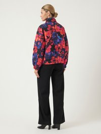 Moteris su šviesiais plaukais su sušukuota kupele, vilkinti spalvingą gėlių rašto siūbtą striukę, juodas plačias kelnes ir juodas aukštakulnius batus, stovi šonu.