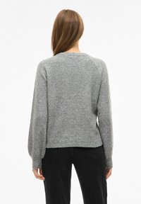 Maglione grigio lavorato a maglia con un motivo texturizzato, scollatura rotonda e polsini a coste. Indossato con pantaloni neri, visto da dietro.