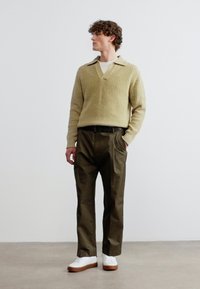 Άνδρας που στέκεται με τα χέρια στις τσέπες, φοράει μπεζ ριγωτό πουλόβερ, σκούρο πράσινο παντελόνι και λευκά αθλητικά παπούτσια, μπροστά σε απλό φόντο.