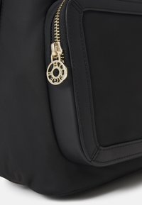 Angolo di una borsa di stoffa nera con zip dorata e tirante circolare che presenta lettere ritagliate sulla piccola tasca frontale.