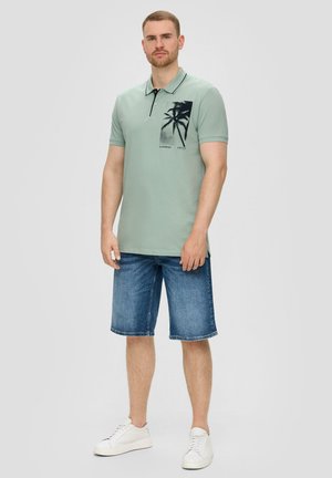 Hellgrünes Poloshirt mit schwarzem Kragen, ausgestattet mit einem Palmbaum-Graphic und kurzen Ärmeln, kombiniert mit Jeansshorts und weißen Sneakern.