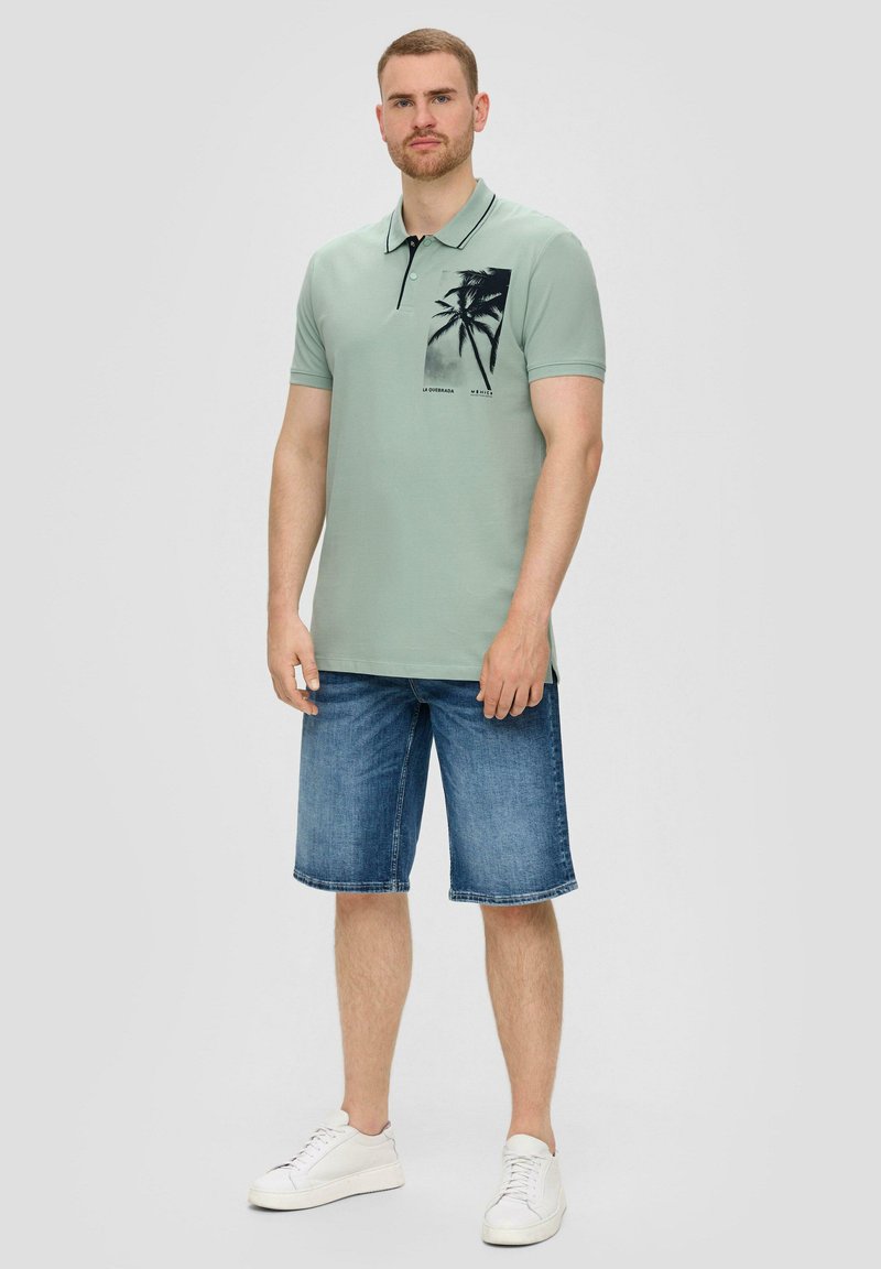 Jasnozielona koszulka polo z czarnym kołnierzykiem, z grafiką palmy i krótkimi rękawami, noszona z dżinsowymi szortami i białymi trampkami.