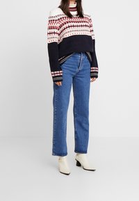 Sweter dzianinowy w kolorze kremowym, granatowym i czerwonym z wzorzystym karczkiem; niebieskie dżinsy z wysokim stanem; kremowe botki do kostki z ostro zakończonym noskiem i słupkowym obcasem.