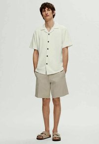 Camisa de botão de manga curta bege claro com botões pretos, combinada com calções caqui claros e sandálias slip-on bege. Design simples, ajuste descontraído.