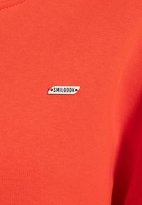 Heldere oranje T-shirt met een gladde textuur en een klein wit label met de tekst "SMILODOX" erop genaaid aan de voorkant.