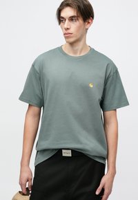 Grå bomulds t-shirt med rund hals, korte ærmer og et lille gult logo på brystet. Kombineret med sorte bukser.