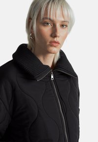 Černá prošívaná bunda s velkým žebrovaným límcem, se stříbrným zipem a hladkou texturou. Detailní pohled, který zdůrazňuje design a švy.