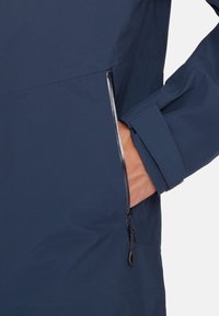 Giacca blu scuro con tessuto testurizzato, tasca laterale con zip e polsini regolabili. Il design è snello e funzionale.