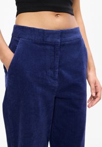 Κοντινό πλάνο ενός ατόμου που φοράει ψηλόμεσα σκούρα μπλε βελούδινα παντελόνια, με το ένα χέρι στην τσέπη και ένα μαύρο κοντό τοπ.