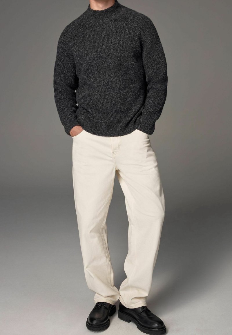 Dunkelgrauer Strickpullover mit hohem Kragen, kombiniert mit cremefarbenen, weit geschnittenen Hosen und schwarzen Schnürschuhen. Glatte Textur und lässige Passform.