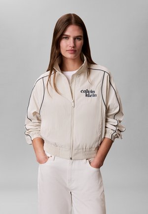 Donna con lunghi capelli castani che indossa una giacca crema Calvin Klein con zip e pantaloni bianchi, in piedi con le mani nelle tasche davanti a uno sfondo grigio.